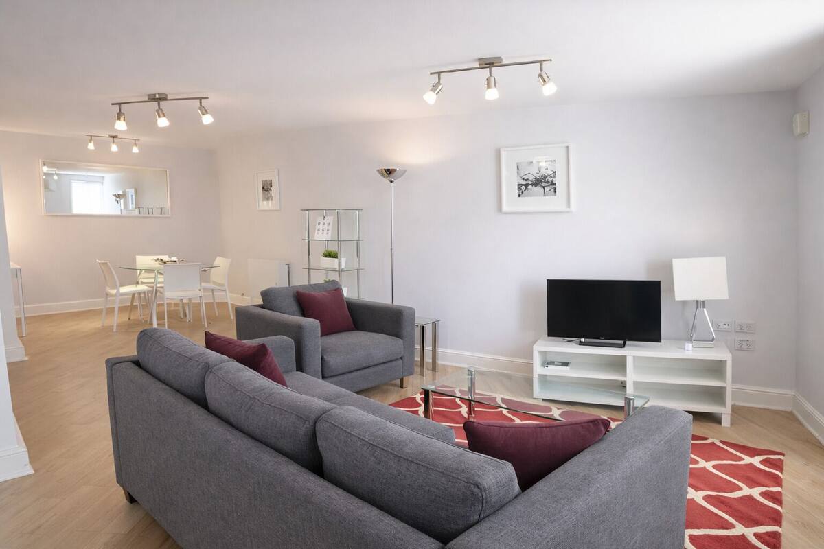 A modern living room with grey sofas, a flat-screen TV, and a dining area with white chairs.