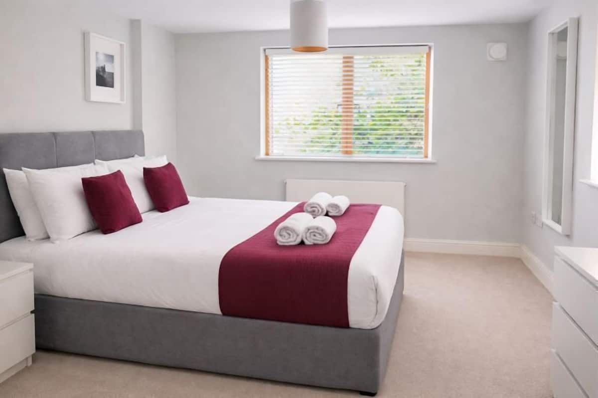 A neatly arranged bedroom with a bed, pillows, a bedside table, and a window with blinds.