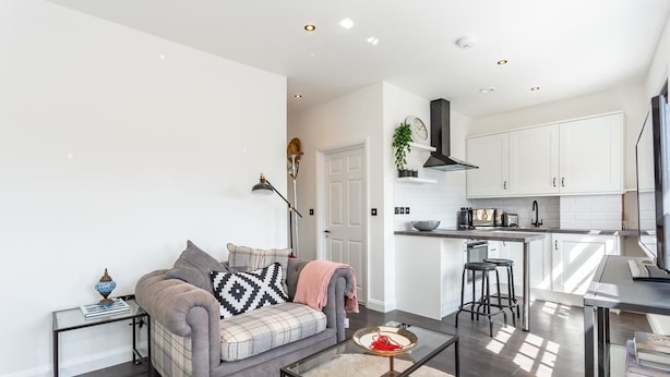 A modern living room with a grey sofa, a coffee table, and a kitchen area with white cabinets and a black range hood. - Namastay in Bed (Southend-on-Sea)
