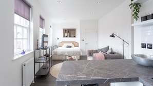 A modern, well-lit room with a bed, a sofa, a dining table, and a kitchen area. - Namastay in Bed (Southend-on-Sea)