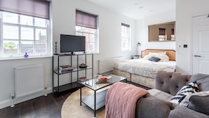 A modern, well-lit room with a bed, a sofa, a TV on a stand, and a window with a view of buildings. - Namastay in Bed (Southend-on-Sea)