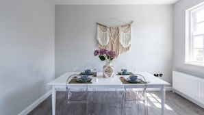 A dining area with a white table, clear chairs, a vase with flowers, and a macrame wall hanging. - Exhale (Southend-on-Sea)