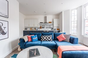 A modern living room with a blue sofa, a coffee table, and a kitchen in the background. - Shape of Water (Southend-on-Sea)