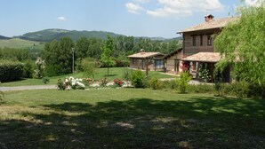 Uma casa de pedra rústica com telhado de telhas, rodeada por um jardim bem cuidado com plantas floridas e uma vista desobstruída de colinas ondulantes ao fundo.