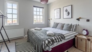 Une chambre à coucher avec un lit, deux fenêtres, un radiateur, une lampe sur pied et une valise.