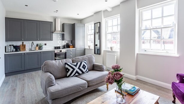 Une cuisine moderne avec des meubles gris, un canapé gris agrémenté d’un coussin noir et blanc, et une table basse en bois sur laquelle est posé un vase de fleurs.