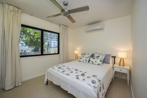 Un dormitorio con cama, dos mesitas de noche con lámparas, un ventilador de techo, un aire acondicionado y una ventana con cortinas.