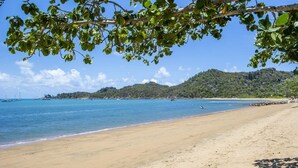 Ein Strand mit klarem, blauem Wasser, sandigem Ufer und einer bewaldeten Hügelkette im Hintergrund.