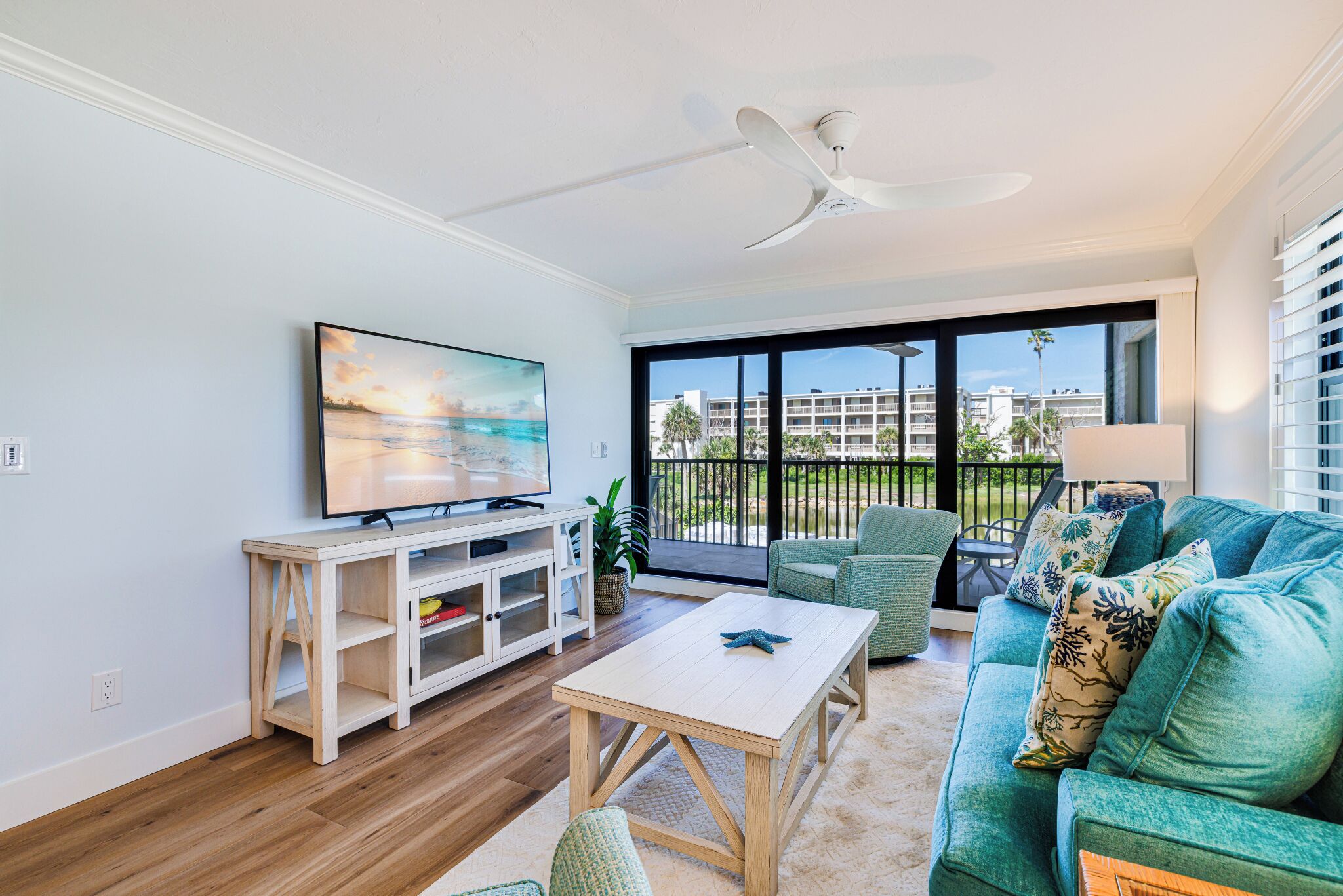 A living room with a TV, a white coffee table, a teal sofa, and a large window with a view of buildings and palm trees.