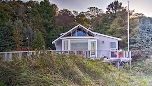 A house with a large bay window, a deck, and a view of the surrounding forest. - Lakefront Montague Cottage w/ Deck & Beach! (Montague)