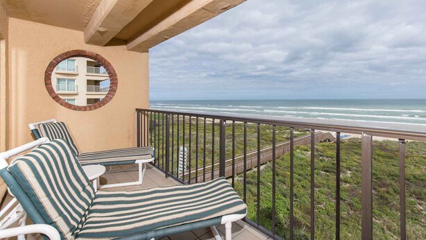A balcony with two striped lounge chairs and a round mirror on the wall, overlooking a beach and ocean. - Suntide II 401 - Beachfront Direct Ocean Views, Private Balcony, Heated Pool, Hot Tub, Beach Access, Walking distance to Dining and Shopping (South Padre Island)