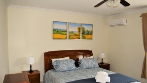 A bedroom with a wooden headboard, two bedside tables with lamps, a bed with a blue quilt, and three landscape paintings on the wall. - Bright Condo steps to Eagle Beach (Oranjestad-West)