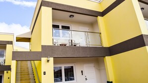 A multi-story building with a yellow facade, a balcony with glass railings, and a staircase leading to the entrance. - Bright Condo steps to Eagle Beach (Oranjestad-West)