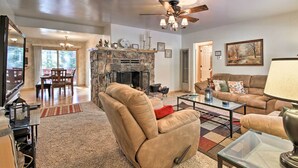 A spacious living room with a stone fireplace, a ceiling fan, and a large window overlooking a deck. - Walk to Lake: Rustic Home w/ Deck in Fawnskin (Fawnskin)