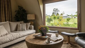A cozy living room with a sofa, a coffee table with a book and a plant, and a view of greenery outside. - Glenlowren - A magical haven in the Yarra Valley (Dixons Creek)