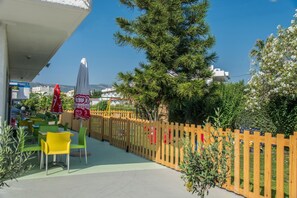 An outdoor seating area with tables and chairs, a wooden fence, and a large tree. - Yannis Yard Studios And Apartments (Kos)
