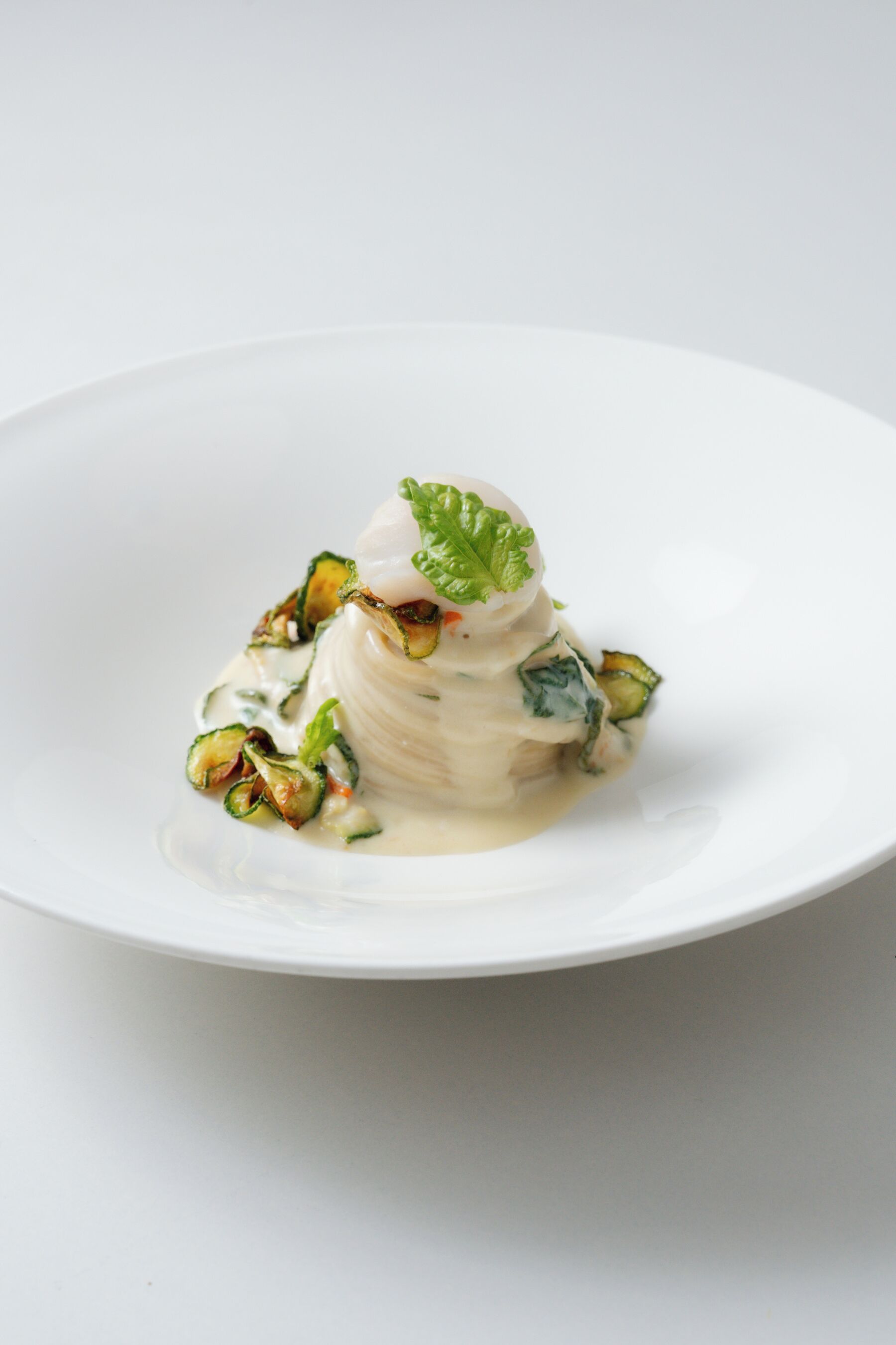 A white plate with a white dessert, possibly panna cotta, topped with a mint leaf and surrounded by a creamy sauce.