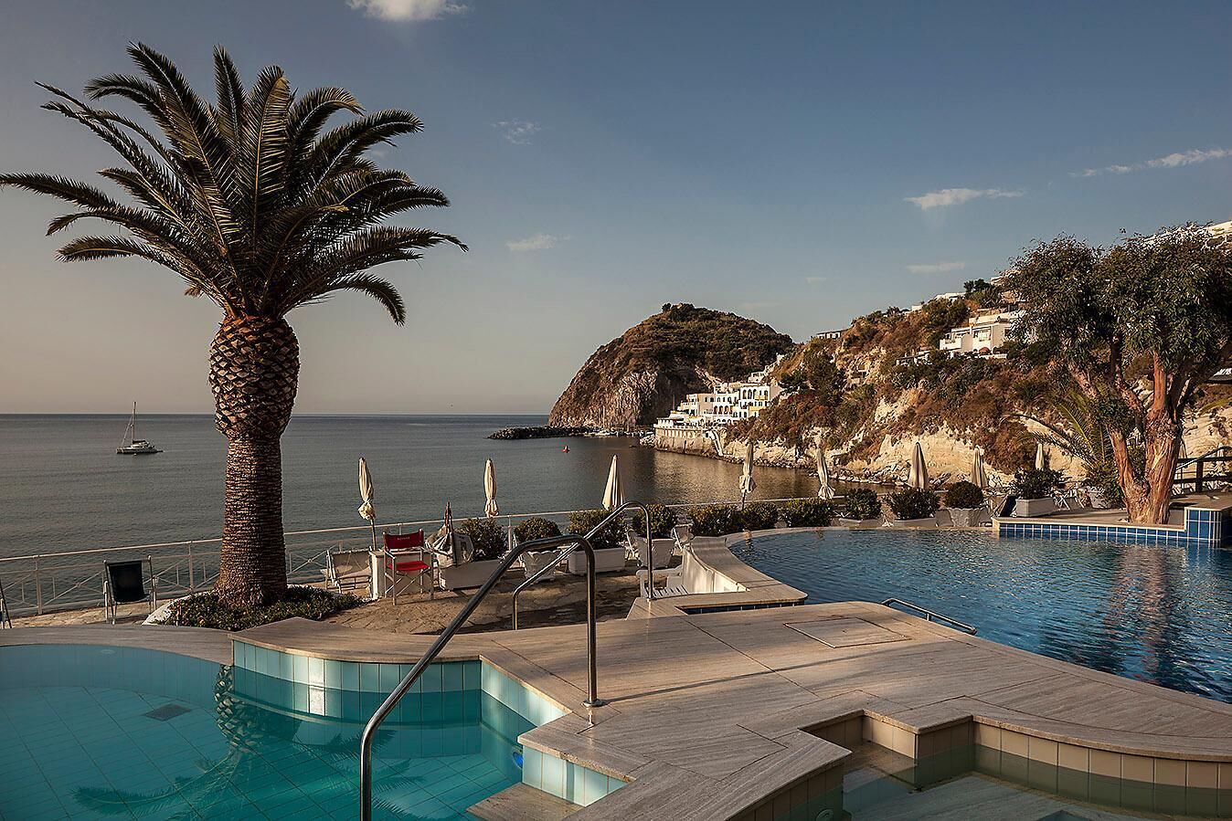 A pool area with a view of the sea, a palm tree, and a mountainous backdrop.
