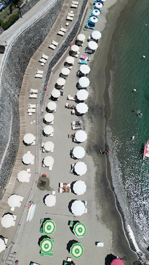 Luchtbeeld van een strand met rijen parasols en ligbedden, een stenen muur en een helder uitzicht op de oceaan.