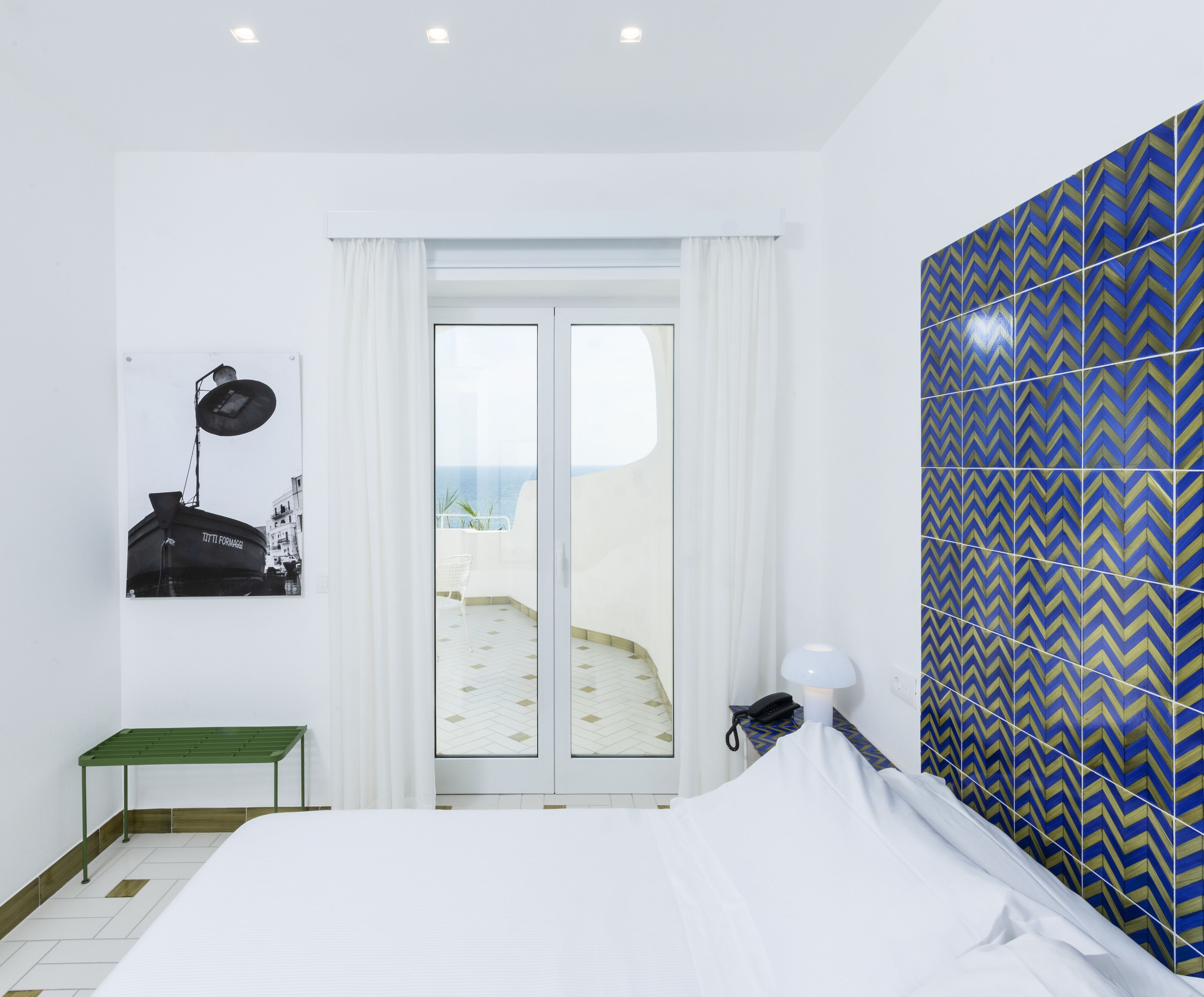 A bedroom with a bed, a bench, a nightstand with a lamp, a phone, and a view of the sea through a glass door.