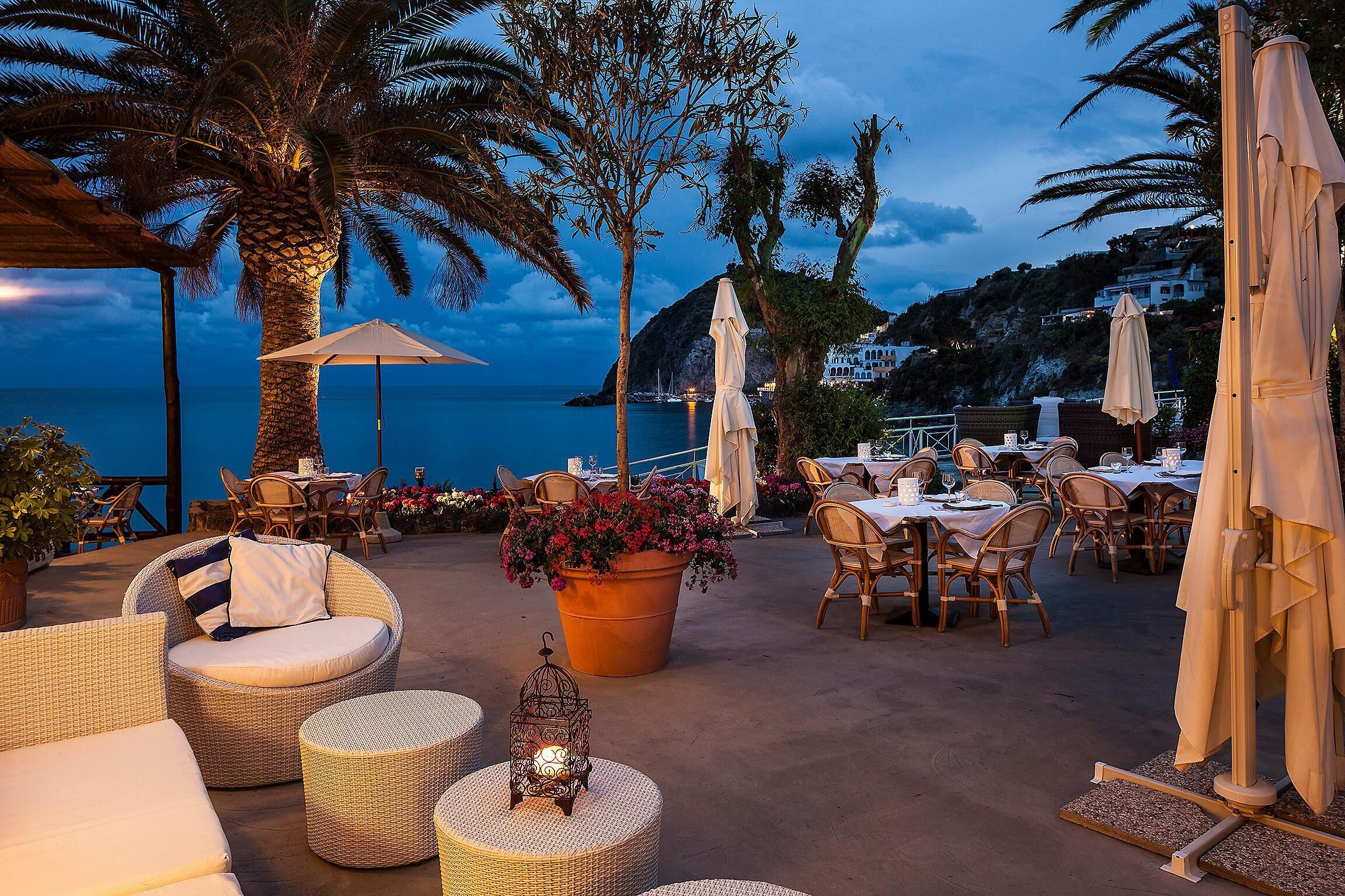 An outdoor dining area with wicker furniture, a large planter with flowers, and a view of the sea at dusk.