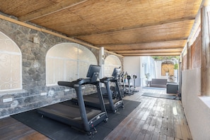 Een overdekte buitenfitnessruimte met stenen muren, houten plafond en trainingsapparatuur, waaronder loopbanden en gewichten.