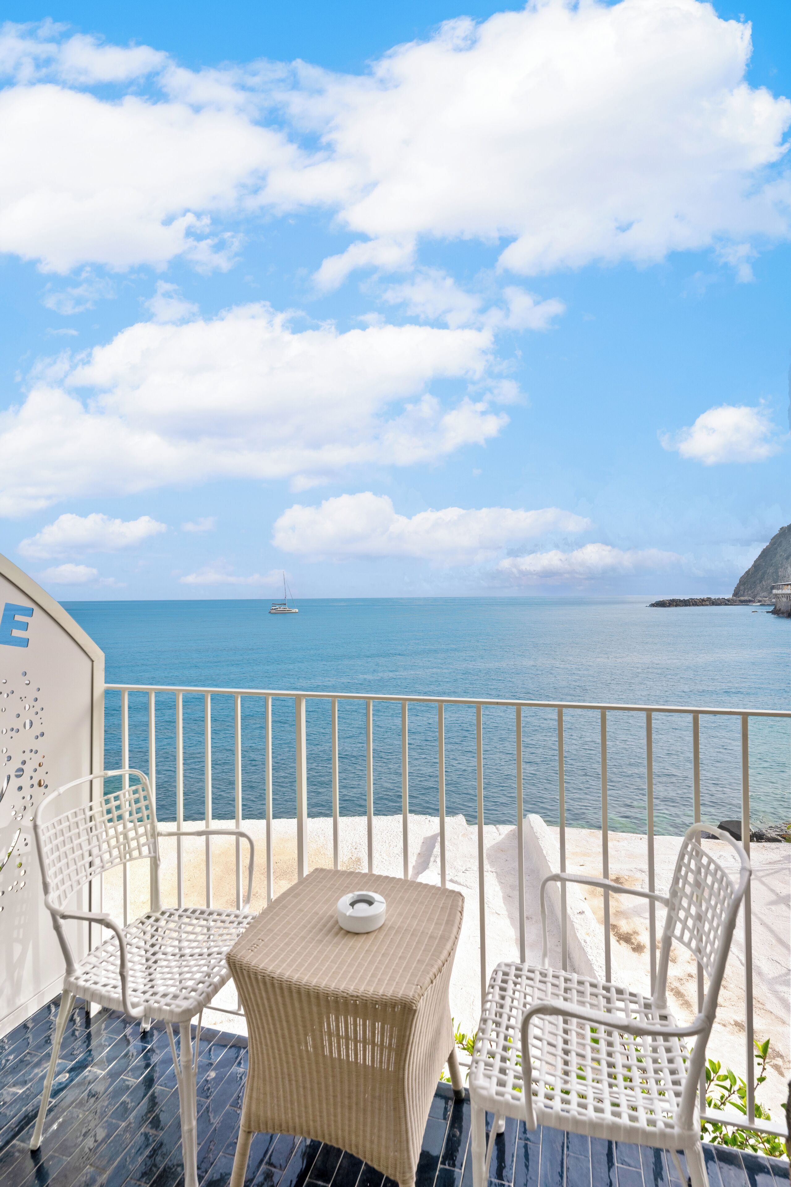 A balcony with two wicker chairs and a small table, overlooking a calm sea with a sailboat in the distance and a rocky coastline.