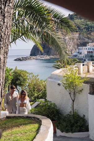 A coastal view with a rocky cliff, a white building with a balcony, and a palm tree.