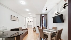 Een moderne woonkamer met een eetgedeelte, een bank, een aan de muur bevestigde televisie en een kroonluchter.