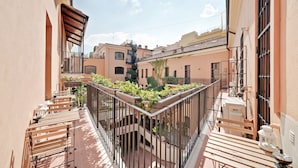 Een smal balkon met houten bankjes en plantenbakken, uitkijkend op een binnenplaats met gebouwen en een trap.