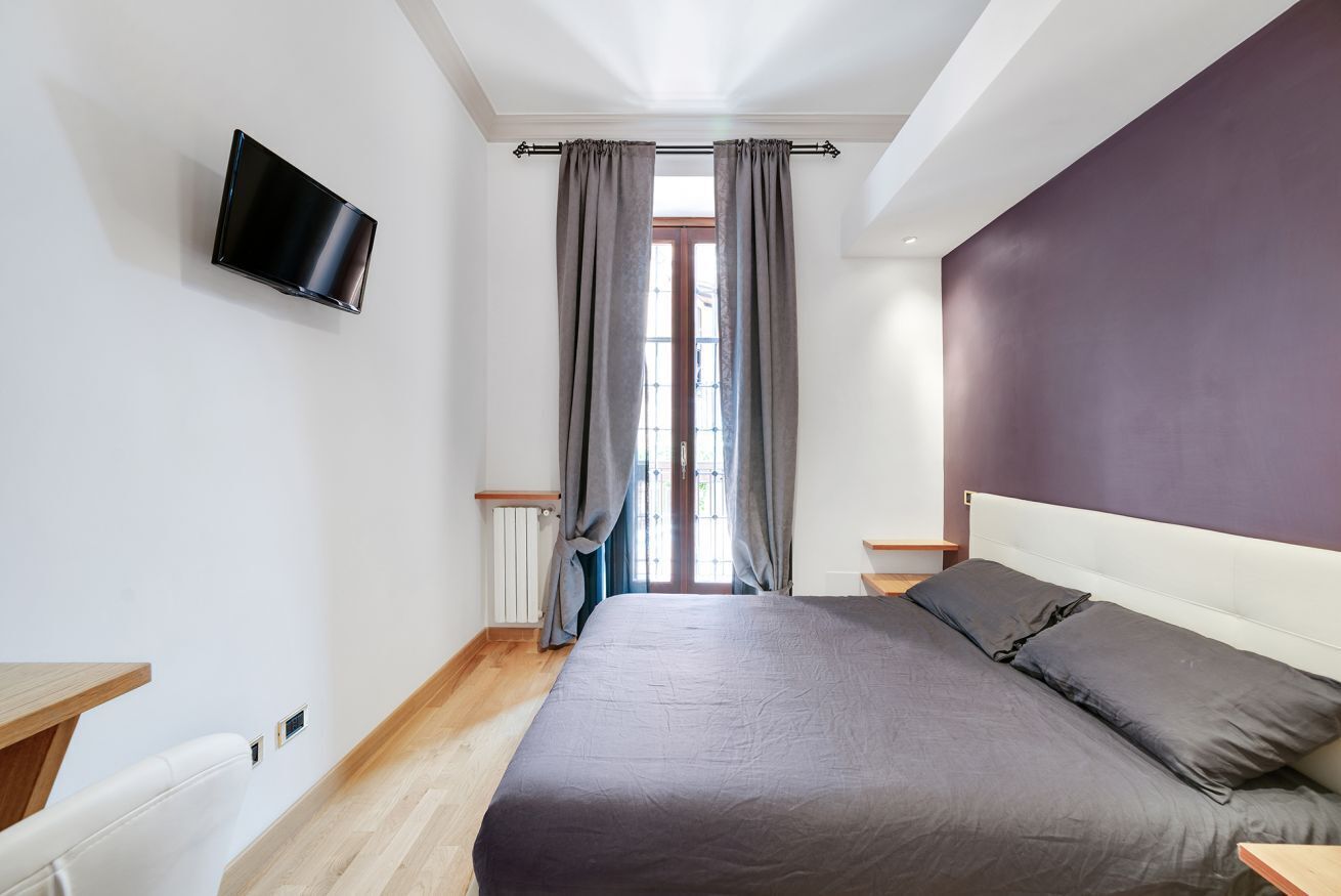 A bedroom with a bed, a TV mounted on the wall, a window with curtains, and a radiator.