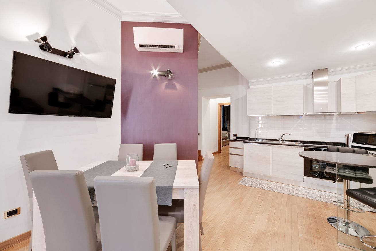 A modern kitchen with white cabinets, a built-in oven, and a dining area with a table and chairs.