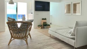 A modern living room with a white sofa, a wicker dining table with striped cushions, a flat-screen TV mounted on the wall, and a view of the ocean through large windows. - Casa Atlantica - beautiful Seaview Apartment (Las Playitas)