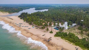 An aerial view of a beach with a row of boats, a swimming pool, and a white building surrounded by palm trees.