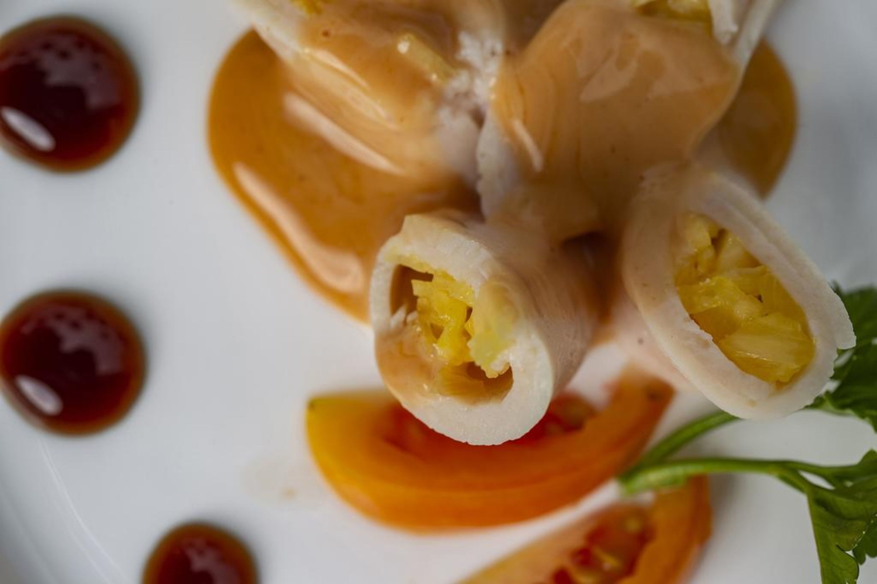 A close-up of a dish with rolled dumplings filled with yellow filling, accompanied by a dark sauce and slices of orange.