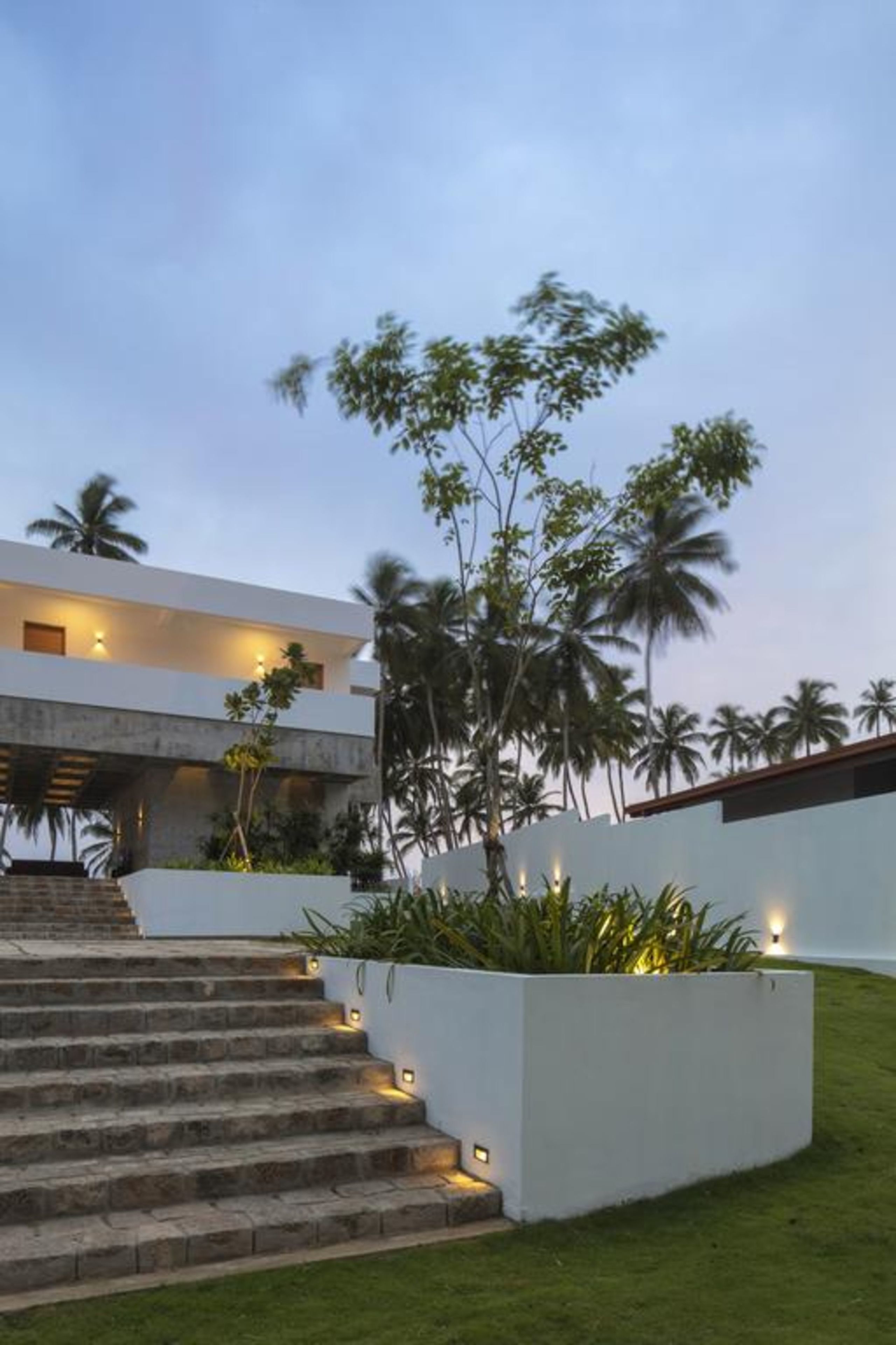 A modern building with a staircase leading to a flat roof, surrounded by greenery and palm trees.
