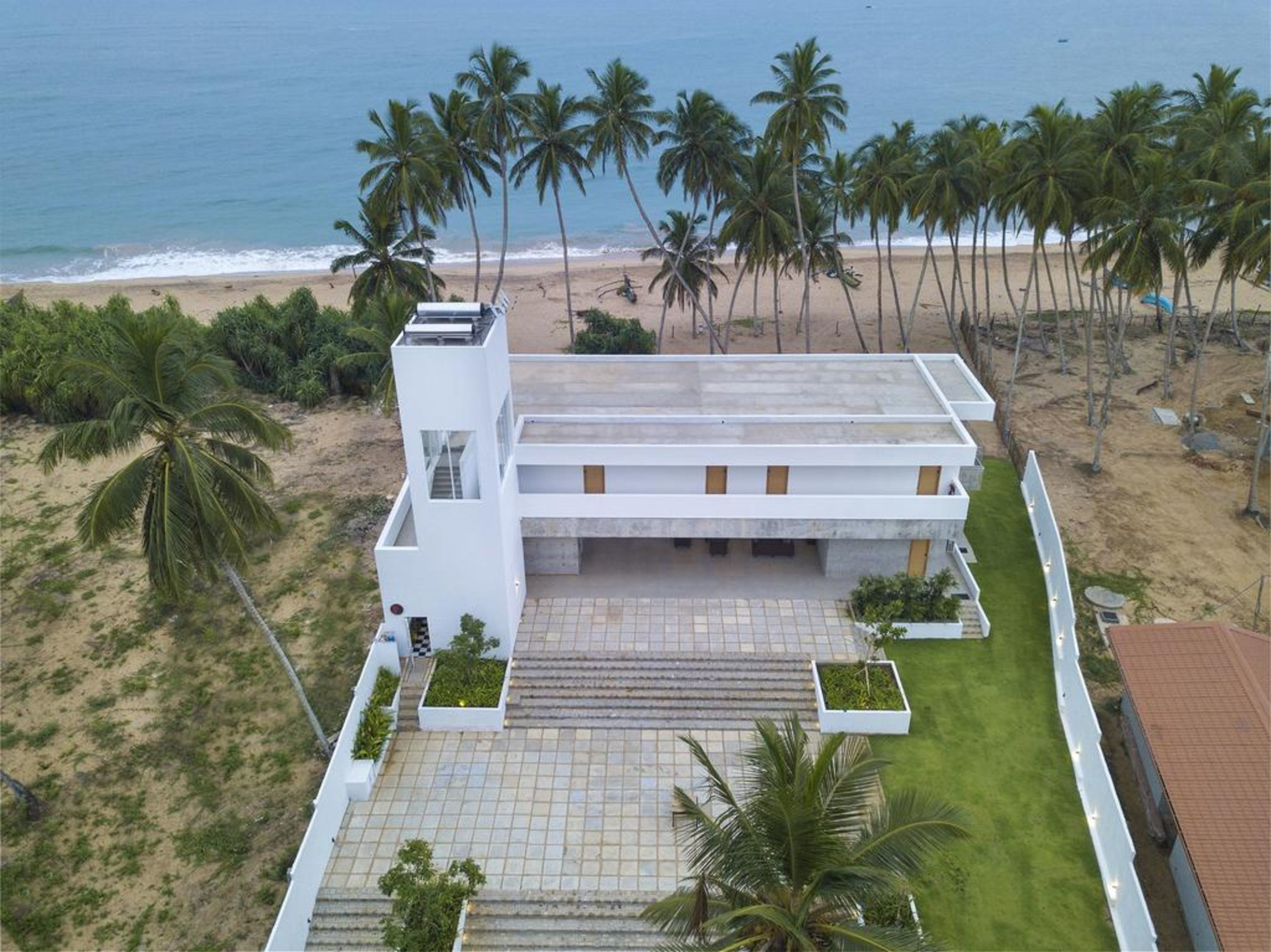 A modern two-story building with a flat roof, surrounded by palm trees and a beach.