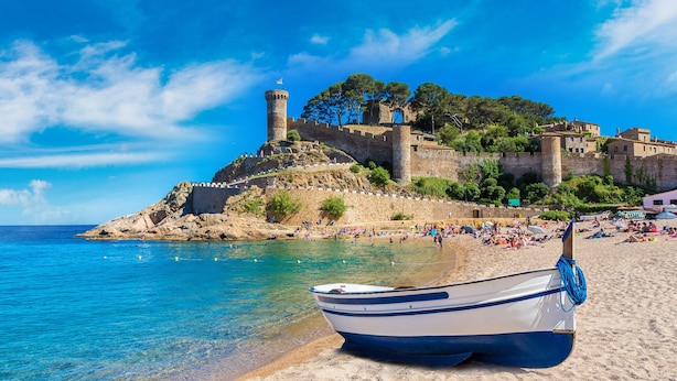 A beach with a boat, clear blue water, and a historic fort on a hill.
