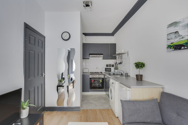 Modern kitchen with central island, oven, microwave, and sink, adjacent to a living area with sofa and flat-screen TV.