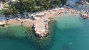 Eine Vogelperspektive zeigt einen Strand mit klarem Wasser, einem steinernen Wellenbrecher und mehreren Liegestühlen und Sonnenschirmen.