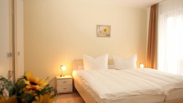 Une chambre d’hôtel avec un grand lit, une table de chevet avec une lampe, un petit tableau accroché au mur et une fenêtre avec des rideaux.