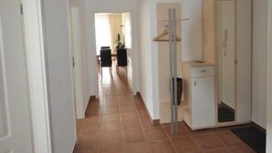 Un couloir avec un sol carrelé, des murs blancs et un luminaire au plafond. Il y a une porte à gauche, un miroir à droite et une petite armoire au milieu.