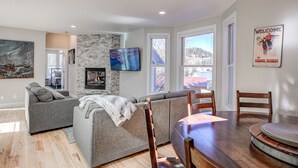 A modern living room with a gray sectional sofa, a wooden dining table with chairs, a flat-screen TV mounted on the wall, and a fireplace.