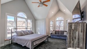 A bedroom with a large bed, a ceiling fan, a TV mounted on the wall, and a view of the outdoors through large windows.