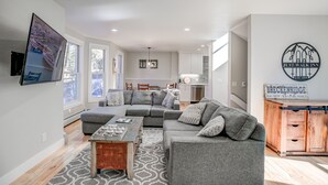 A modern living room with a gray sectional sofa, a wooden coffee table, and a patterned rug. There is a kitchen area with white cabinets and a dining table. A sign on the wall reads "Just Walk Inn."