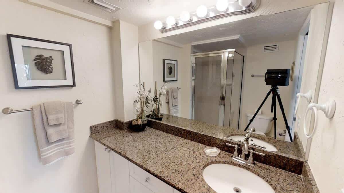 A bathroom with a granite countertop, two sinks, a large mirror, and a camera on a tripod.
