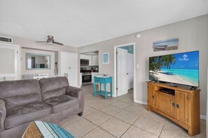A living room with a grey sofa, a television on a wooden cabinet, and a kitchen in the background. - Beachfront Unit! Gulf View from Every Room. Lanai (Siesta Key)
