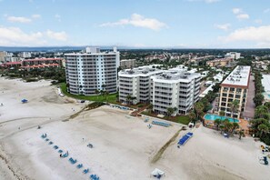 A beachfront hotel with multiple high-rise buildings, a sandy beach with blue lounge chairs, and a swimming pool. - Beach & Pool Open! Beachfront View Every Room (Siesta Key)