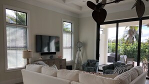 Una sala de estar con un sofá blanco grande, un televisor de pantalla plana, una lámpara con trípode y vistas al exterior a través de amplias ventanas.