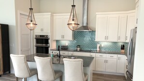 Una cocina moderna con gabinetes blancos, encimera de granito, una isla central con taburetes de bar, electrodomésticos de acero inoxidable y salpicadero de azulejos azules.